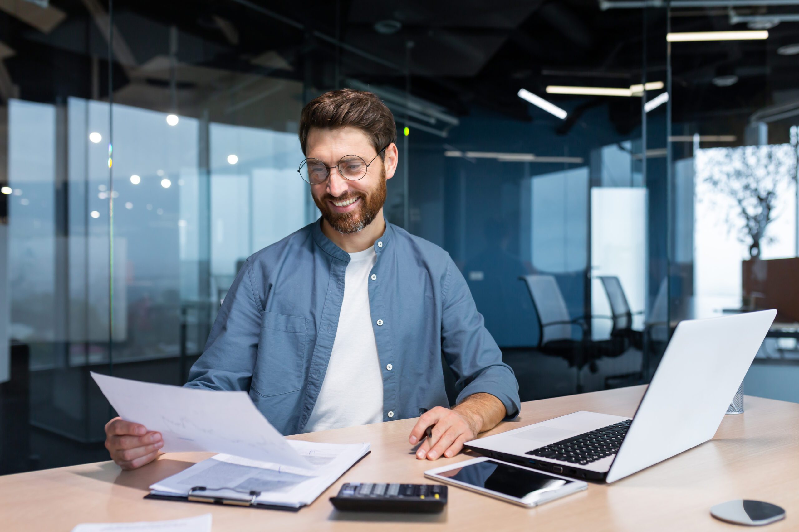 Successful financier investor works with documents and accounts inside office, businessman with beard smiling satisfied with work results and achievements, man inside office with laptop working